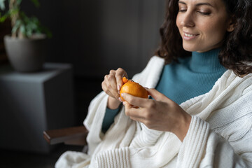 Woman with fresh fragrant tangerine at home. Selective focus of happy cheerful female covered with plaid peeling juicy citrus fruit while relaxing indoors, eating vitamin C rich food in winter season