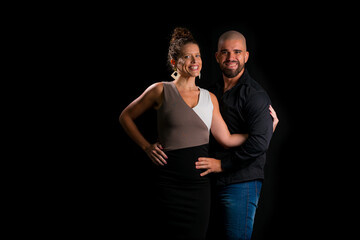 Obraz premium Retrato de casal jovem em estúdio com fundo preto. Mulher loira vestindo vestido social e homem branco careca vestindo camisa social preta e jeans. Abraçados olhando para a câmenta.