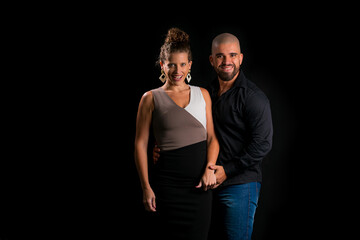 Obraz premium Retrato de casal jovem em estúdio com fundo preto. Mulher loira vestindo vestido social e homem branco careca vestindo camisa social preta e jeans. Homem e mulher sorrindo para a câmera.
