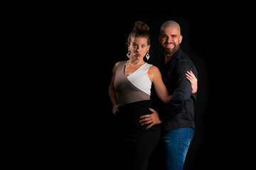 Obraz premium Retrato de casal jovem em estúdio com fundo preto. Mulher loira vestindo vestido social e homem branco careca vestindo camisa social preta e jeans. Mulher fazendo careta e homem sorrindo.