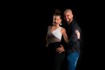 Retrato de casal jovem em estúdio com fundo preto. Mulher loira vestindo vestido social e homem branco careca vestindo camisa social preta e jeans. Modelos fazendo careta e sorrindo.