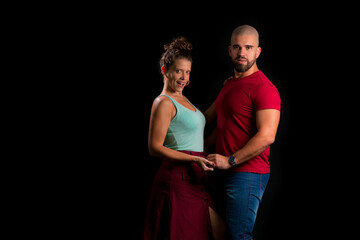 Retrato de casal jovem em estúdio com fundo preto. Mulher loira vestindo blusa verde vestido vermelho e homem branco careca vestindo blusa vermelha e bermuda jeans. Olhando para a camera de mãos dadas