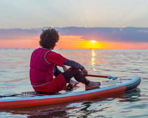 A young man enjoys the sunrise over the sea sitting on a paddleboard.