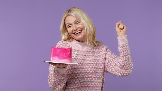 Excited happy fun elderly blonde woman lady 40s years old wears warm shirt look camera hold birthday cake with candle dance clench fist isolated on plain pastel light purple background studio portrait