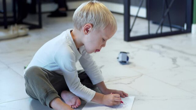 A blonde caucasian 4 years old boy is drawing a picture with colorful pencils on the floor at home and showing his art into the camera. Domestic concept