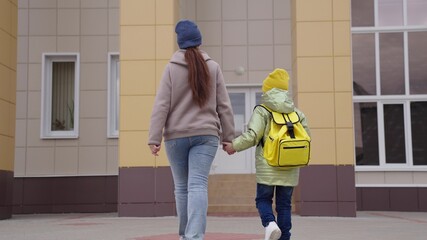 mother leads little kid with school backpack to school, boy, an elementary school student hurries to lesson in class, elementary education of kid teenager, girl with bag is holding mother by the hand