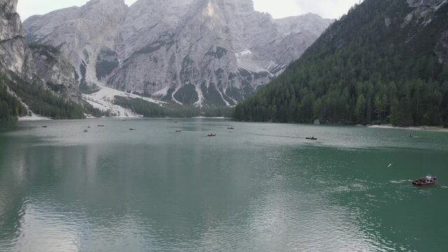 Drone video with a diagonal plane, over the lake of braies with boats with people rowing and mountains on the horizon.