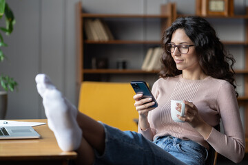 Young relaxed female employee holding smartphone and tea cup resting at workplace, selective focus....