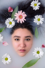 Obraz na płótnie Canvas Closeup portrait of the face of a beautiful young woman in a milk bath self care spa surrounded by delicate white and pink flowers