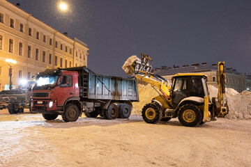 Snow removal equipment working at Palace Square at night after heavy snowfall. Snow cyclone or blizzard paralyzed city, municipal tractors load collected snow into dump truck. Winter season concept. 