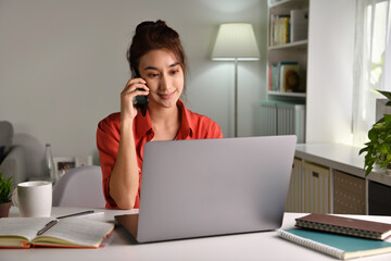 Beautiful young  Asian woman work at home with laptop and talking on smartphone and smiling. Female working on computer and using cell mobile phone while sitting at home