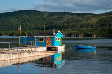 A small boathouse at the end of a wooden pier. The bright teal blue building has a red door and...