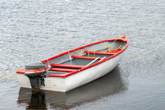 A white wooden traditional dory or small fishing vessel with red trim sits on a smooth water surface. The dory has wooden oars and rope.  The smooth water is reflecting the image of the boat. 