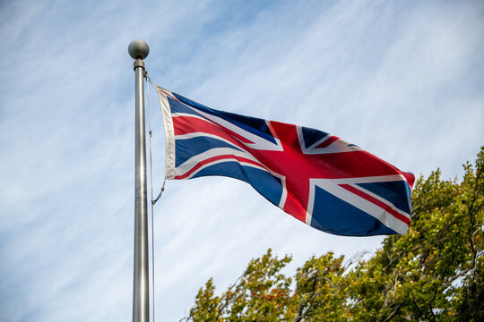 The European flag is known as the Union Jack flying on a metal pole with blue sky and clouds in the background. The red, white, and blue flag is actually three crosses representing the United Kingdom.