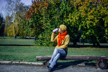 Pretty european woman in yellow jacket and red scarf sitting on wooden bench in a deserted park and drinking coffee. Enjoying solitude and sunny autumn weather. Blurred plant background, copy space.