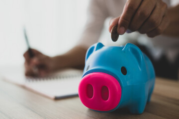 A young employee is putting coins into a blue piggy bank and recording income and expenses on the...