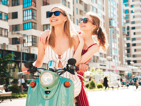 Two young beautiful smiling hipster female in trendy overalls.Sexy carefree women driving retro motorbike on the street background.Positive models having fun, riding classic Italian scooter in eyewear