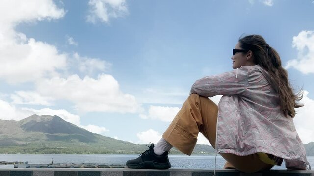 A girl with long dark hair wearing sunglasses, a yellow pants is sitting on shore of lake with volcano. Female traveler sits on shore of pond with green mountain, her clothes fluttering in the wind.