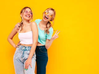 Fototapeta Two young beautiful smiling blond hipster female in trendy summer clothes. Sexy carefree women posing near yellow wall in studio. Positive models having fun. Cheerful and happy. In sunglasses obraz
