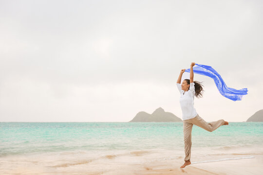 Freedom on beach happy carefree Asian woman jumping free in the wind with blue scarf running during ummer vacation. Positivity, well-being in your body, health and wellness for women.