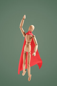 Brave Wooden Man In Red Cloak Fly Up With Raised Right Hand On Green Backgrond. Minimal Concept Of Heroism.