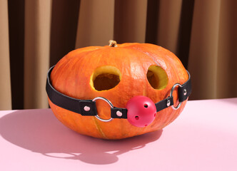 Round big orange pumpkin with eyes cut out and red ball gag on pink table. Halloween Party and...