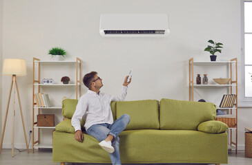 Man resting on sofa at home turns on wall air conditioner pressing power switch on remote control. Happy guy relaxing on comfortable sofa in white Scandinavian Nordic interior with good AC system