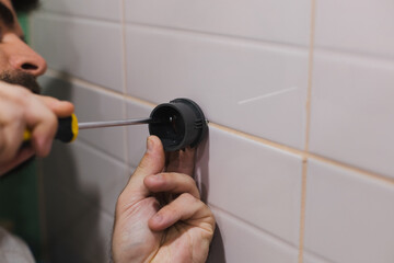 Man hands attaching plastic component of shower head holder to tiled wall in washroom with screwdriver with part of face in background. Renovation works in bathroom. Dirt from renovation process.