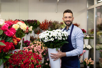 a man florist in the refrigerator among fresh flowers holding a vase with live plants in his hands looks at the camera with a smile
