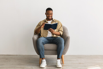 Black Man Reading Book Sitting In Armchair On Gray Background