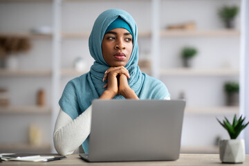 Thoughtful Black Muslim Freelancer Woman In Hijab Sitting At Desk With Laptop