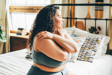 Curly haired overweight young woman in grey top and shorts with satisfaction on face accepts curvy body shape standing in stylish bedroom closeup - obrazy, fototapety, plakaty