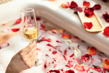 Female hand holding a glass of white wine while sitting in a bathtub with foam and red rose petals. Enjoying life concept. Selective focus