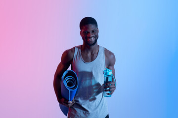 Yoga for healthy living. Happy young black guy holding sports mat and bottle of water in neon light