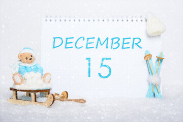 December 15th. Teddy bear sitting on a sled, blue skis and a calendar date on white snow. Day 15 of month.