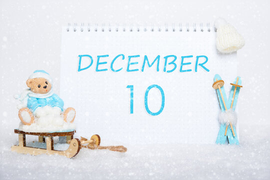 December 10th. Teddy bear sitting on a sled, blue skis and a calendar date on white snow. Day 10 of month.