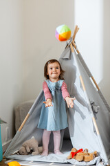 childhood, hygge and people concept - little baby girl playing with ball in kid's tent or teepee at...