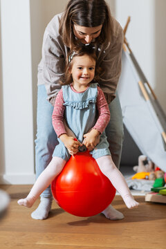 family, leisure and people concept - happy mother playing with little baby daughter bouncing on hopper ball at home