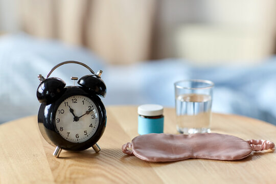 sleep disorder, bedtime and morning concept - close up of alarm clock, eye sleeping mask, glass of water and soporific medicine on night table at home