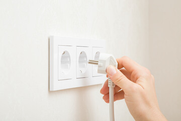 Young adult woman hand holding and plugging white electrical plug in wall outlet socket at home. Closeup. Side view. - obrazy, fototapety, plakaty