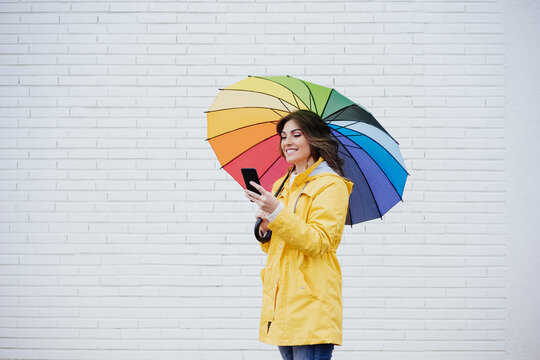 Woman with umbrella using mobile phone in front of wall