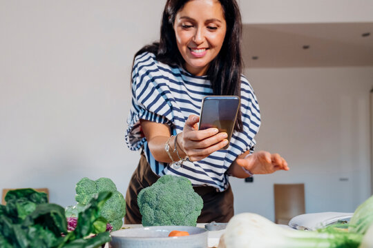 Smiling woman taking picture of fresh vegetables on table at home