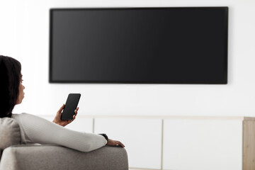 Modern gadgets. Black lady with smartphone connected to TV, watching video at home, plasma...