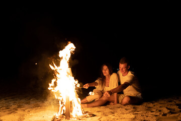 A young couple on the seashore sits by the fire and toasts marshmallows on a stick. A romantic date by the fire. Marshmallow kebab. Tourism and travel concept. - Powered by Adobe