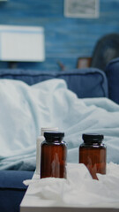 Ill man sleeping on couch with blanket while having cold and flu. Person resting because of virus disease and having bottles of pills, medicaments, cup of tea and tissues on table.
