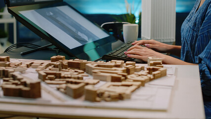 Close up of building model on table and woman using touch screen, analyzing maquette on computer. Architect working on construction plan and layout for architectural development project