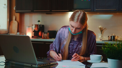 Woman in pajama and with medical mask signing documents. She working at the computer in the kitchen at home.