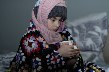 person in warm clothes sitting on a bed with cup of tea