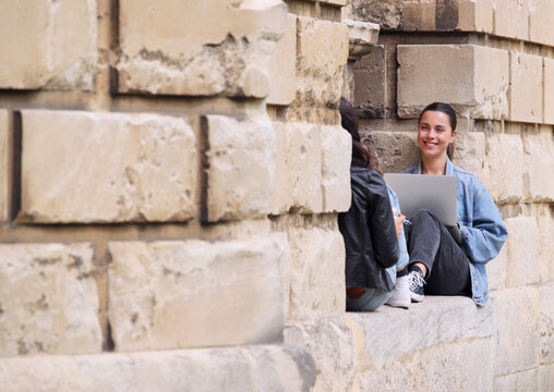 Two Female University Or College Students Sitting Against Wall In City Working On Digital Tablet