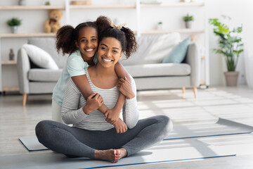 Sporty black mother and daughter sitting on fitness mat, embracing